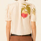 a cream-colored short-sleeve blouse by Alix of Bohemia with a tie-neck detail and colorful embroidered fruit motifs
