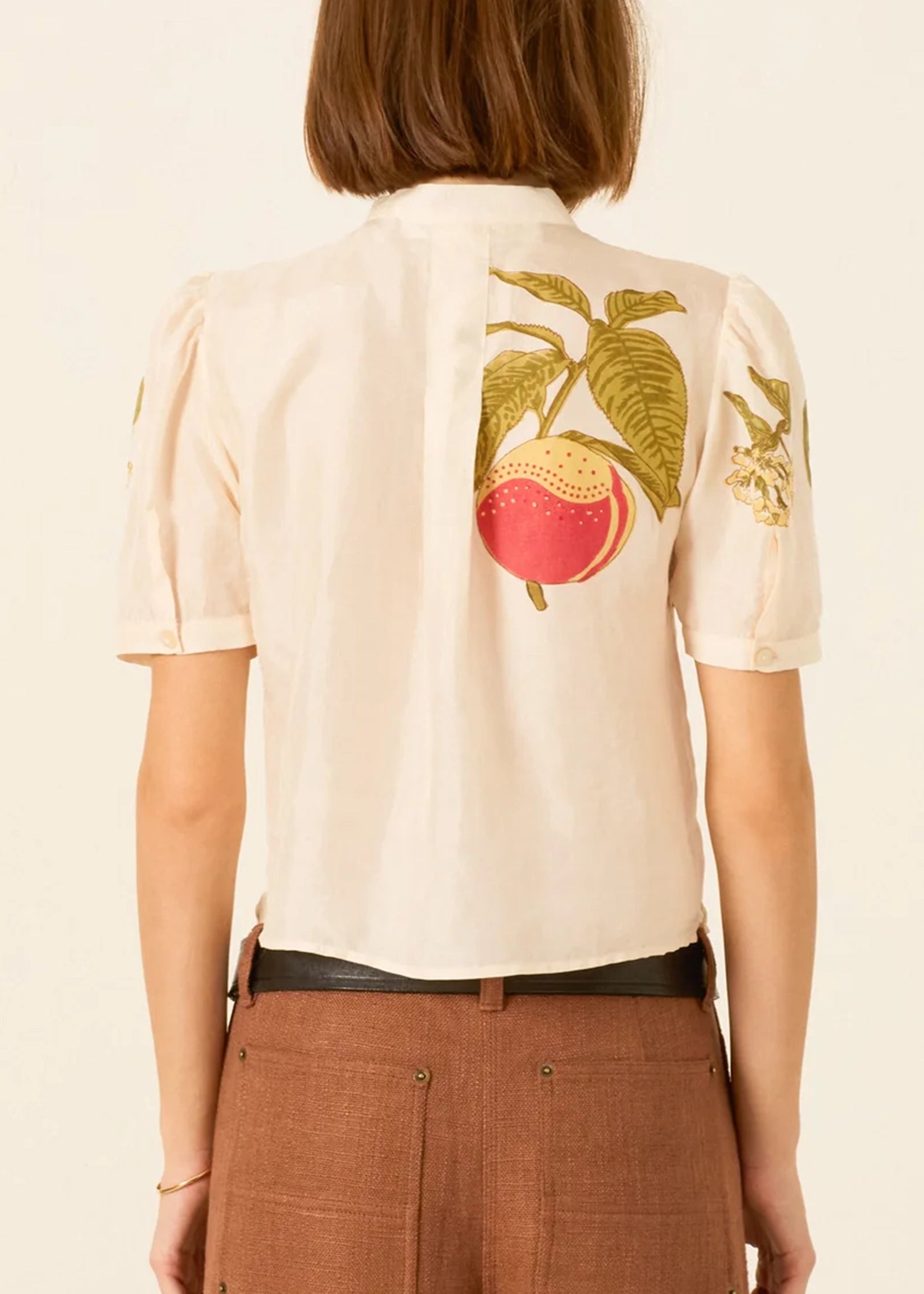 a cream-colored short-sleeve blouse by Alix of Bohemia with a tie-neck detail and colorful embroidered fruit motifs